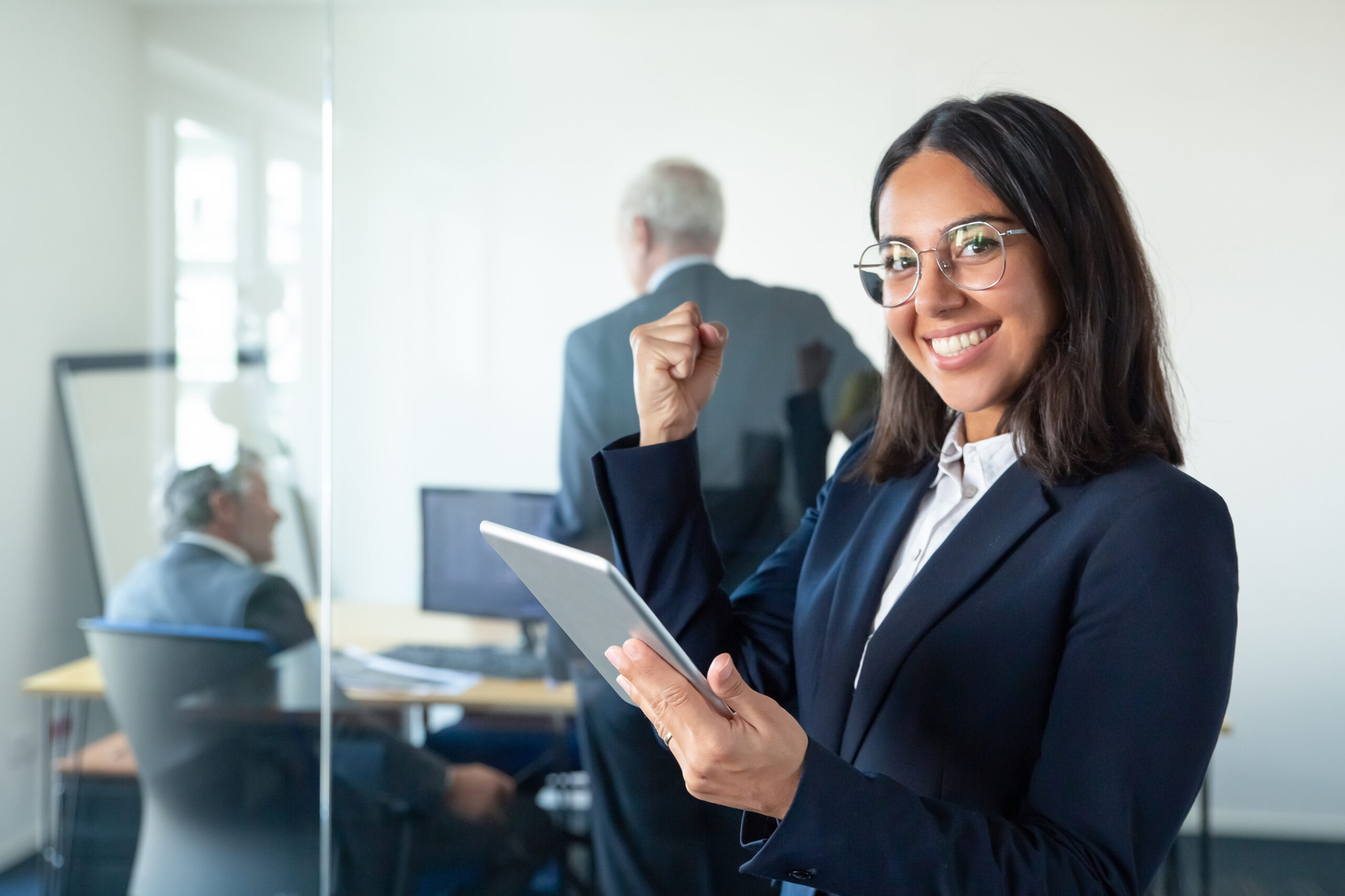 Happy female professional in glasses and suit holding tablet and making winner gesture while two businessmen working behind glass wall. Copy space. Communication concept